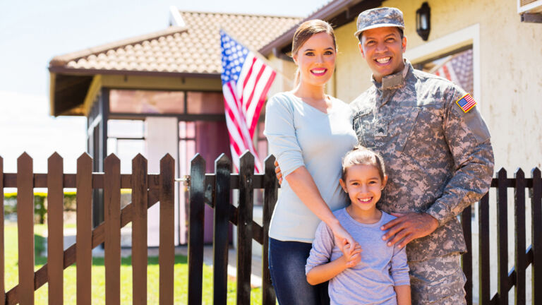 A family, wife and military husband with their daughter in front of picket fence with American flag behind them