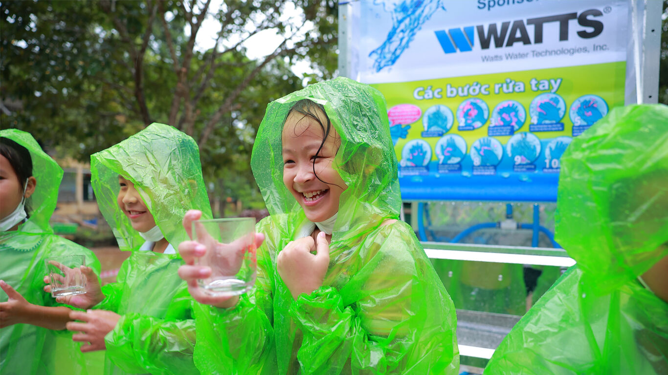 Children wearing green ponchos in the rain standing in front of Watts water tower holding cups of water and smiling.