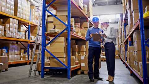 Man and woman standing together in warehouse looking over a clipboard discussing. 